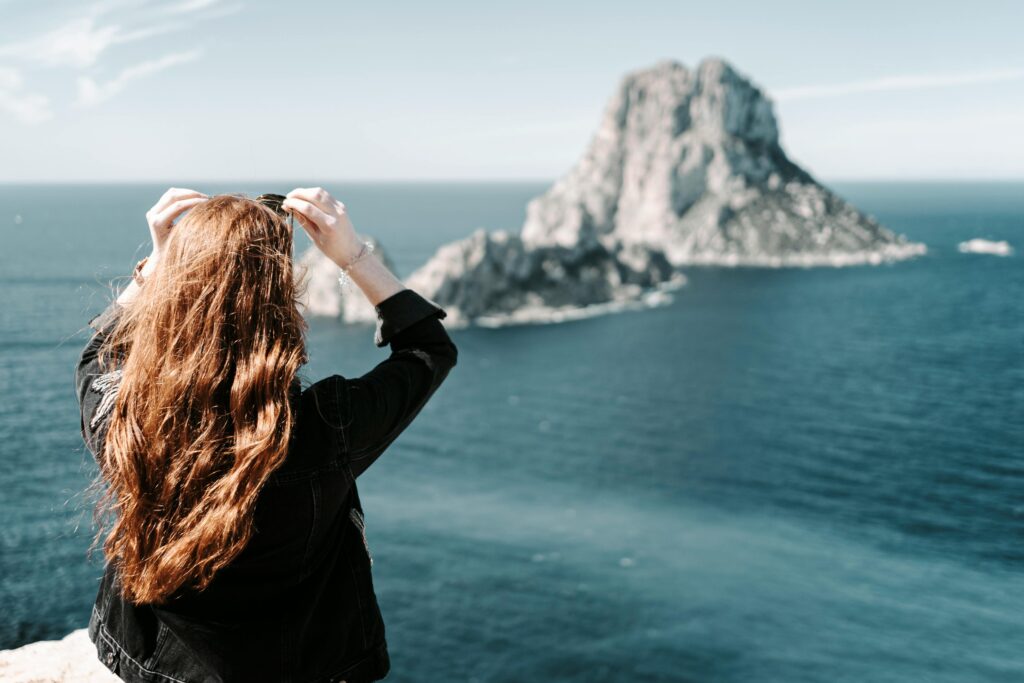 A woman photographs the stunning Es Vedrà island from a cliff in Ibiza under a clear sky.